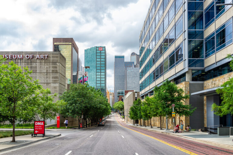 City street leading toward downtown Dallas, with the Dallas Museum of Art on the left, modern office buildings on both sides, and trees lining the roadway.