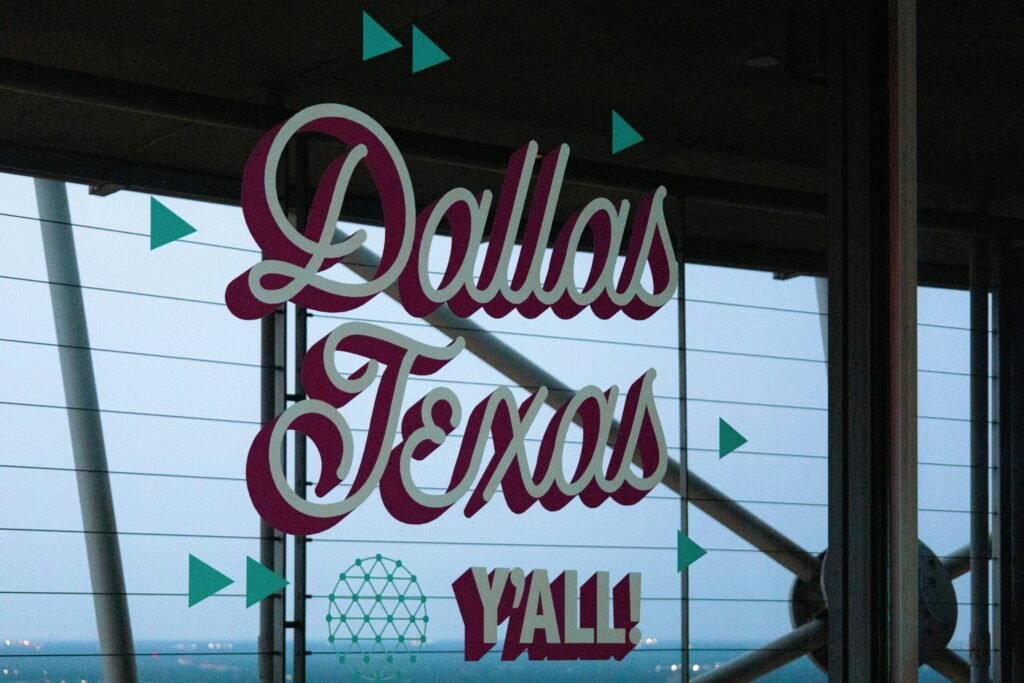 Neon sign reading “Dallas, Texas” displayed inside a building with windows and decorative geometric accents.