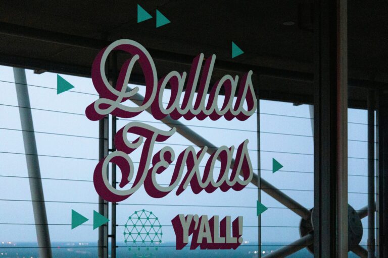 Neon sign reading “Dallas, Texas” displayed inside a building with windows and decorative geometric accents.
