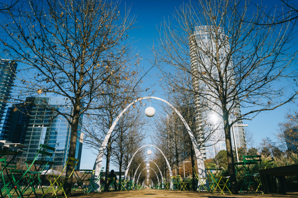 Tree-lined urban walkway with arch structures and green outdoor chairs