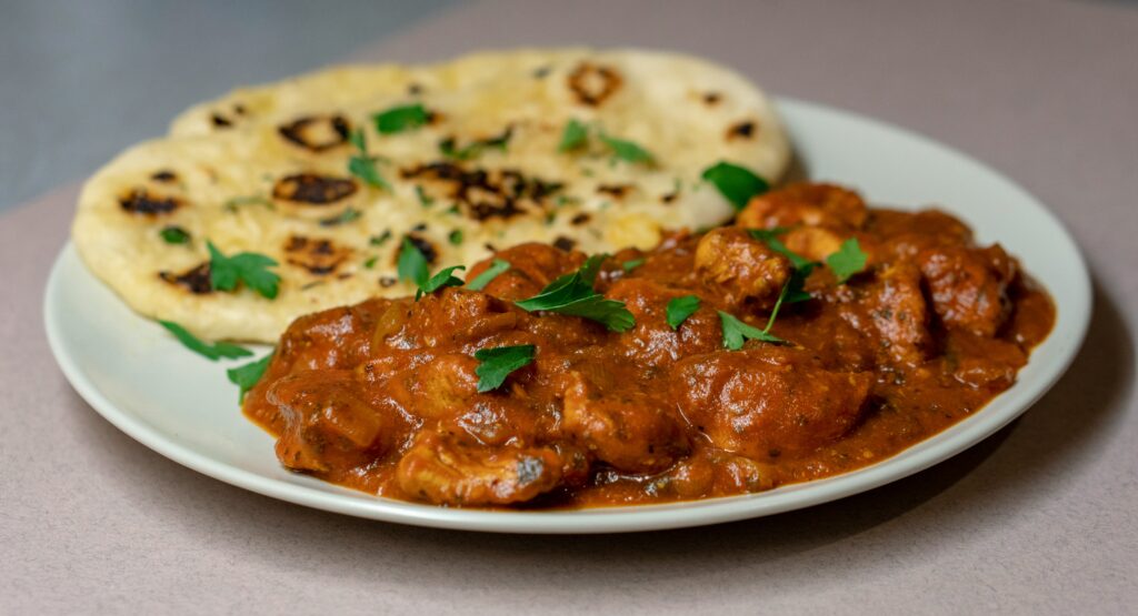 Plate of Indian curry with tender pieces of meat in a rich sauce, served alongside naan bread garnished with herbs.