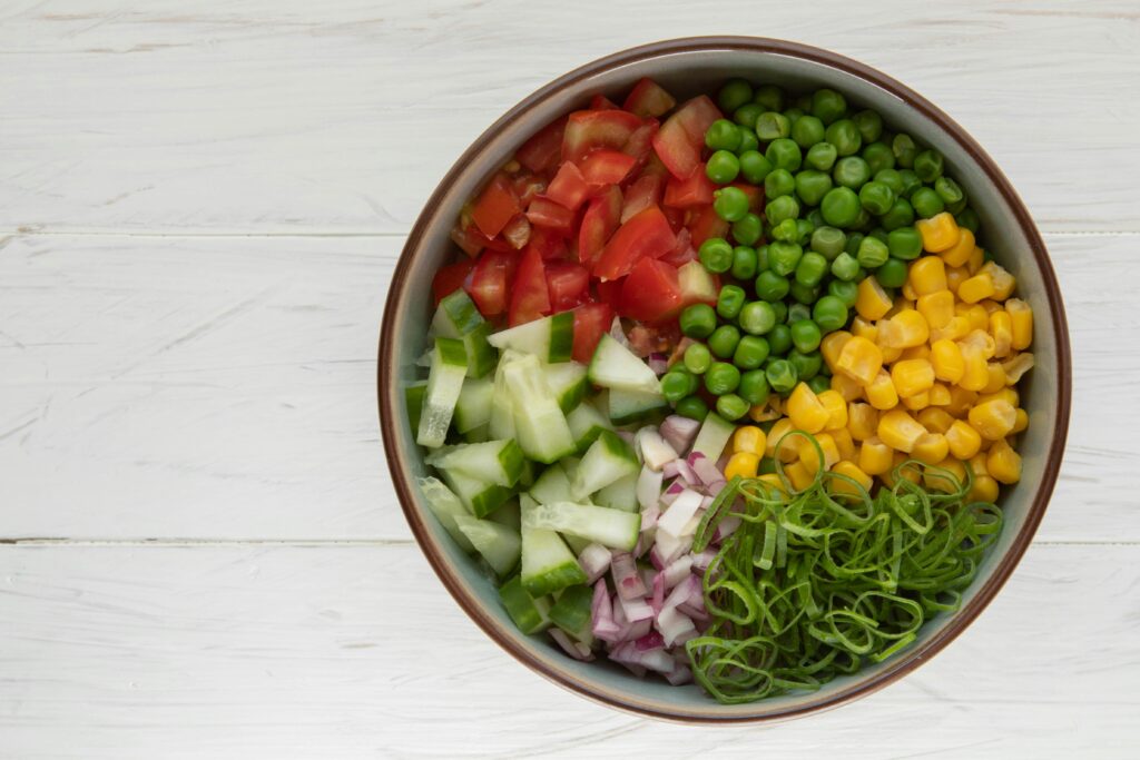 Bowl of chopped vegetables including tomatoes, cucumbers, peas, corn, red onion, and green herbs arranged in sections.