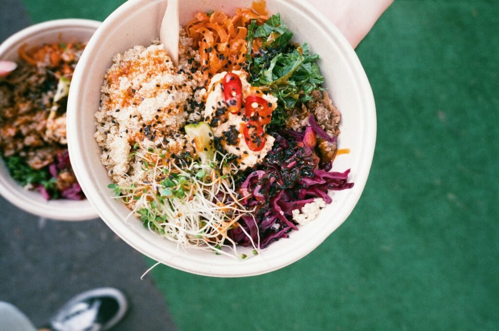 Hand holding a bowl filled with quinoa, mixed vegetables, shredded greens, sprouts, and sauce, photographed from above.