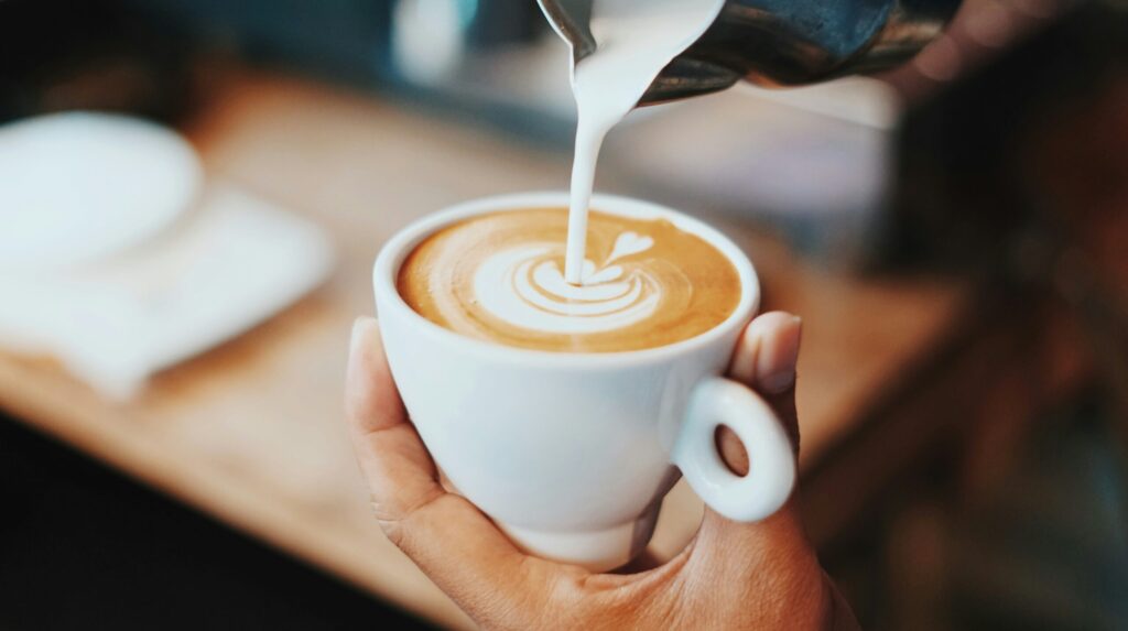 Barista pouring steamed milk into a cup of coffee to create latte art, with the mug held in one hand over a wooden counter.