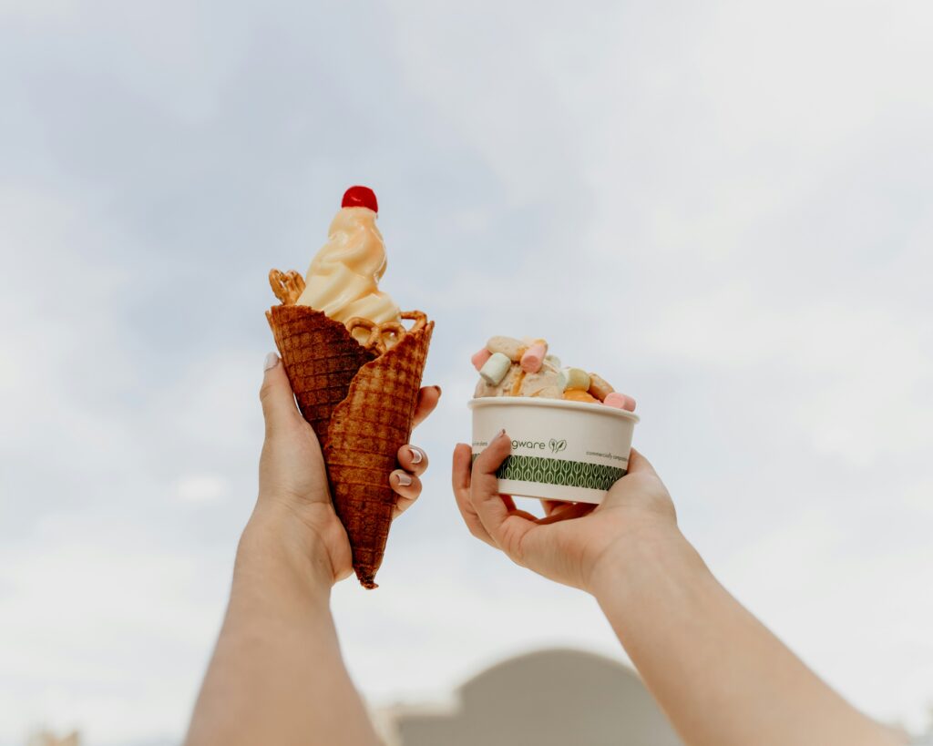 Two hands holding ice cream desserts against the sky, one with a soft-serve cone topped with a cherry and the other with a cup of ice cream and marshmallows.