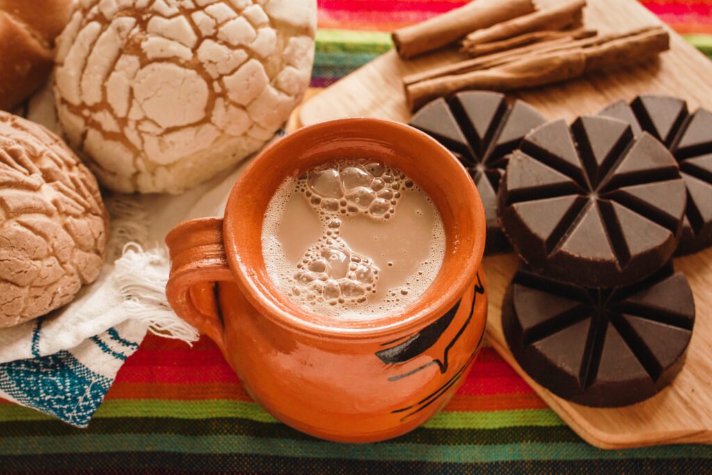 Traditional Mexican hot chocolate served in a clay mug, surrounded by pan dulce bread and a wooden molinillo on a colorful tablecloth.