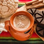 Traditional Mexican hot chocolate served in a clay mug, surrounded by pan dulce bread and a wooden molinillo on a colorful tablecloth.