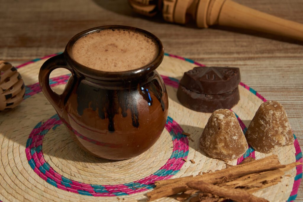 Clay mug of traditional Mexican hot chocolate on a woven mat, surrounded by cinnamon sticks, piloncillo cones, chocolate, and a wooden molinillo.