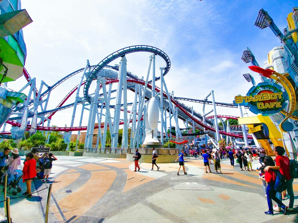 Amusement park walkway with visitors walking beneath a looping roller coaster track, colorful buildings, and a Starbot Café sign.