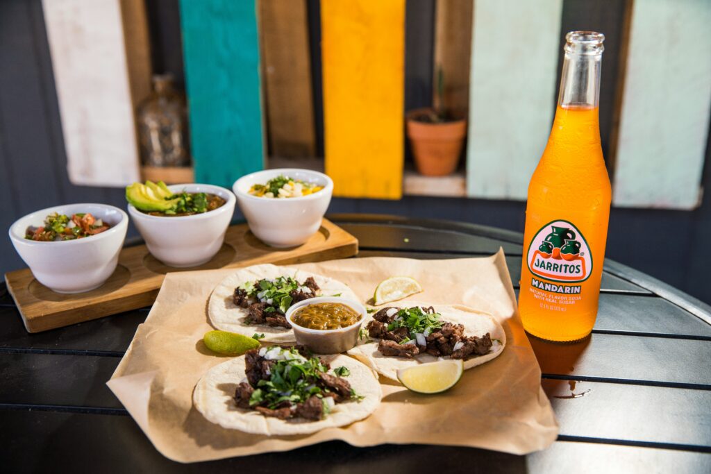 Street-style tacos topped with chopped meat and cilantro on a paper-lined table, served with lime wedges, salsas, and a bottle of Mandarin Jarritos soda.