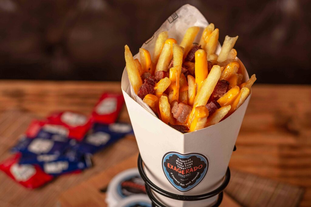 Cone of French fries topped with melted cheese and bacon pieces, served in a branded paper holder on a wooden table.