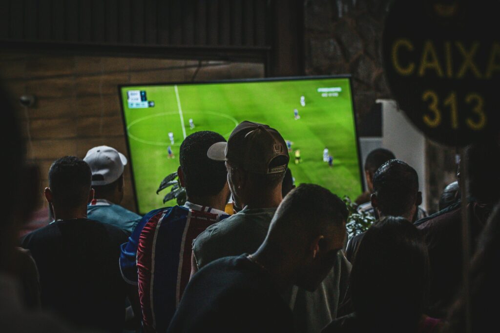 Group of people standing closely together in a bar, watching a soccer match on a television screen.