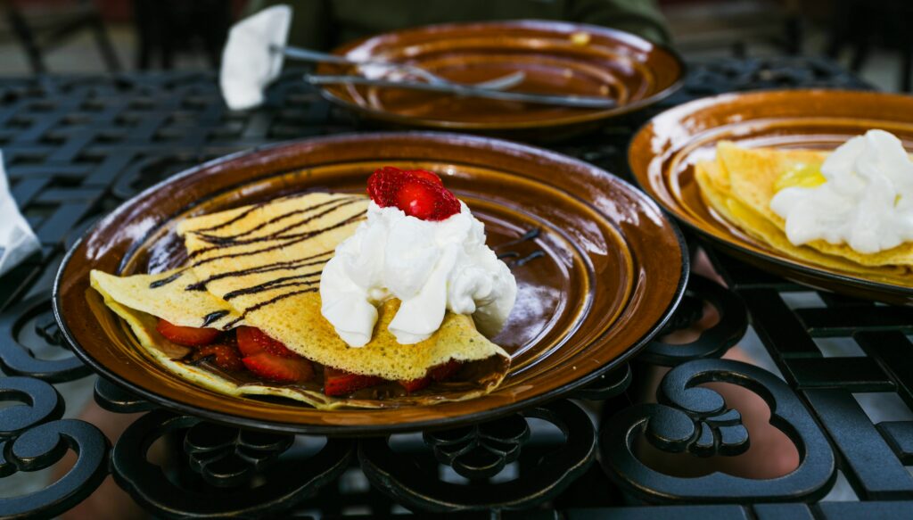 Plates of sweet crepes filled with fruit and topped with whipped cream, served on a decorative outdoor table.