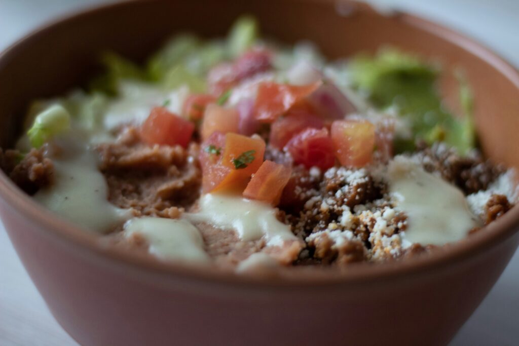 Bowl of Mexican-style food with seasoned meat, lettuce, pico de gallo, crumbled cheese, and creamy sauce.