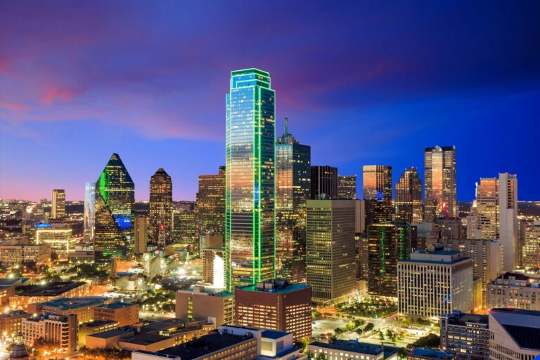 Dallas skyline at dusk with illuminated skyscrapers, including a tall tower outlined in green lights, against a colorful evening sky.