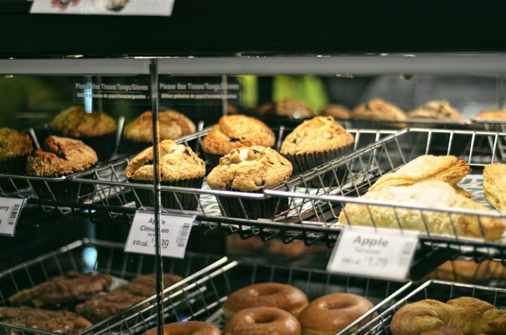 Bakery display case with rows of muffins, pastries, and donuts arranged on metal racks with price labels.