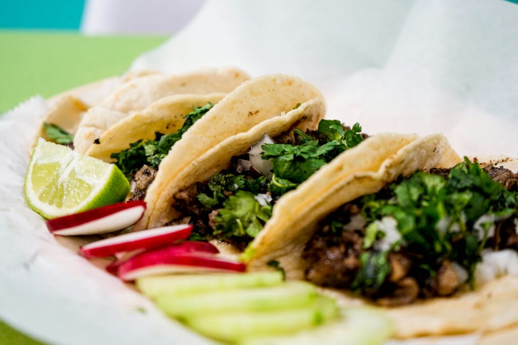 Three street tacos filled with chopped meat and cilantro in soft corn tortillas, served with lime wedges, sliced radishes, and cucumber.