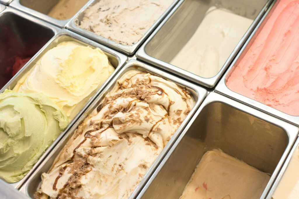 Assorted ice cream flavors displayed in metal tubs, including vanilla, chocolate swirl, pistachio, and strawberry.