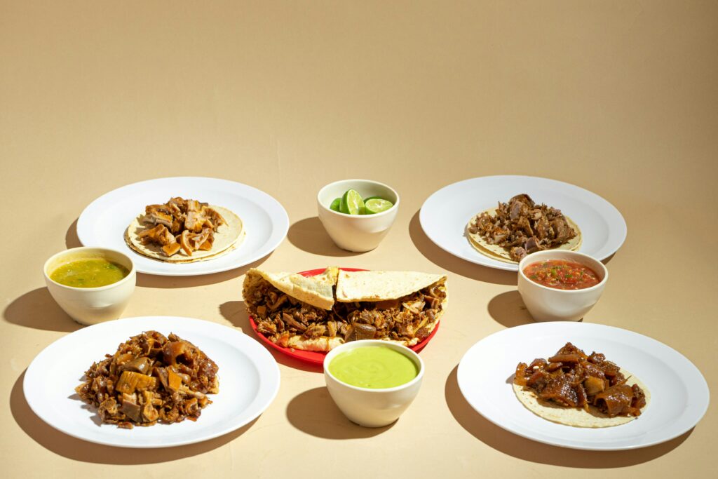 Plates of tacos filled with shredded meat arranged on a table, with small bowls of green and red salsa and lime wedges.