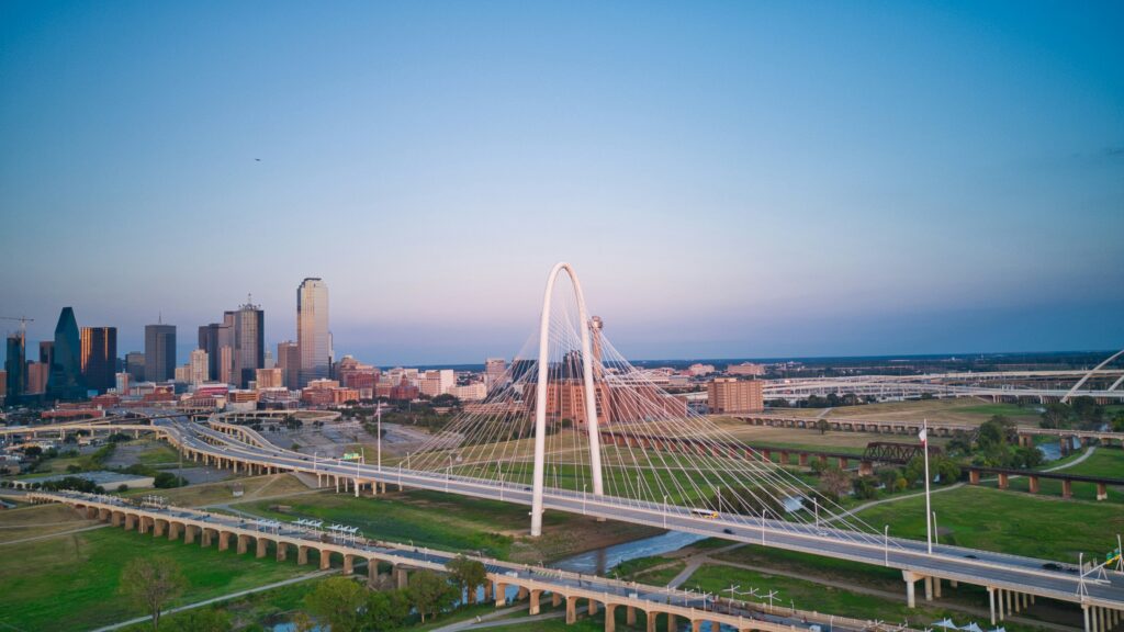 Aerial view of a modern white arch bridge spanning a river, with the Dallas skyline visible in the background under a clear blue sky.