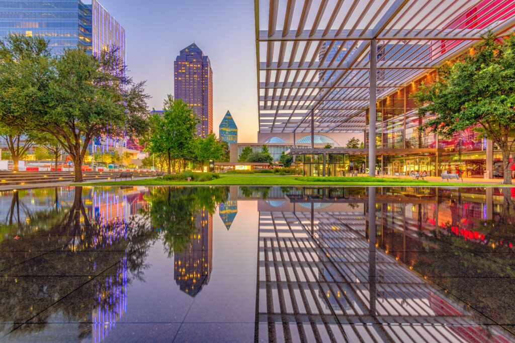 City skyline at dusk reflected in a shallow water feature, with modern office towers, trees, and a covered walkway with metal slats in an urban plaza.