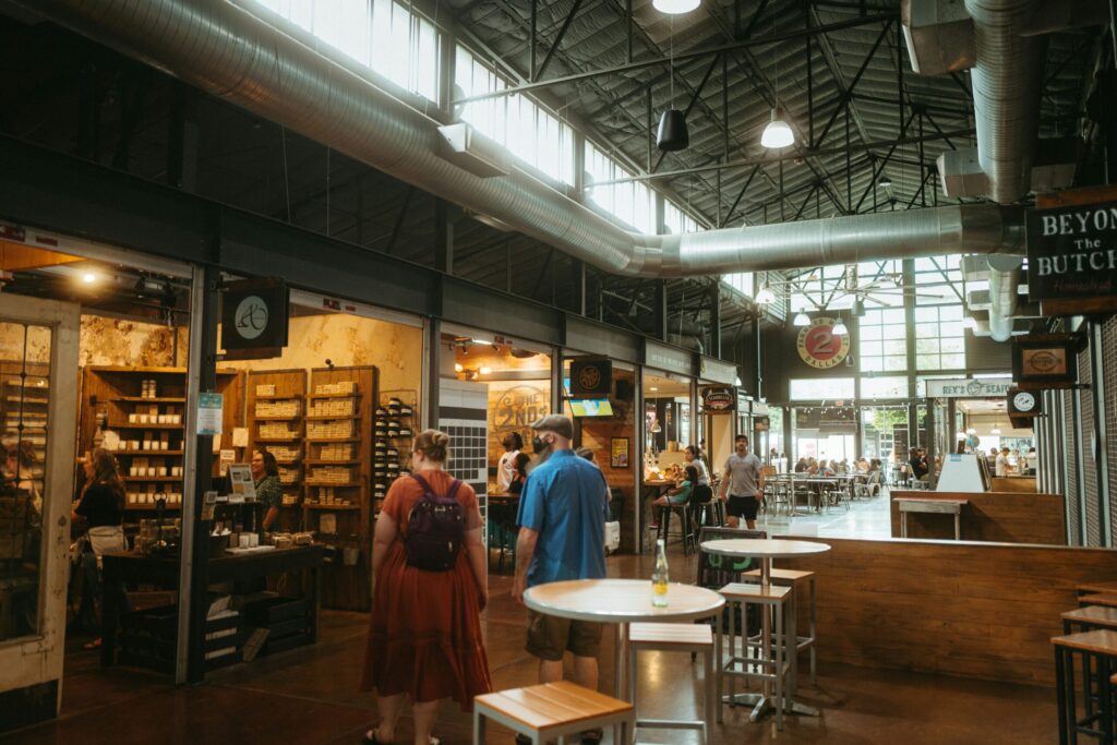 Indoor food hall with high industrial ceiling, exposed ductwork, market-style food stalls, and people walking between shared tables and vendor counters.