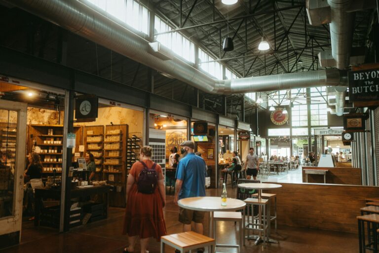 Indoor food hall with high industrial ceiling, exposed ductwork, market-style food stalls, and people walking between shared tables and vendor counters.