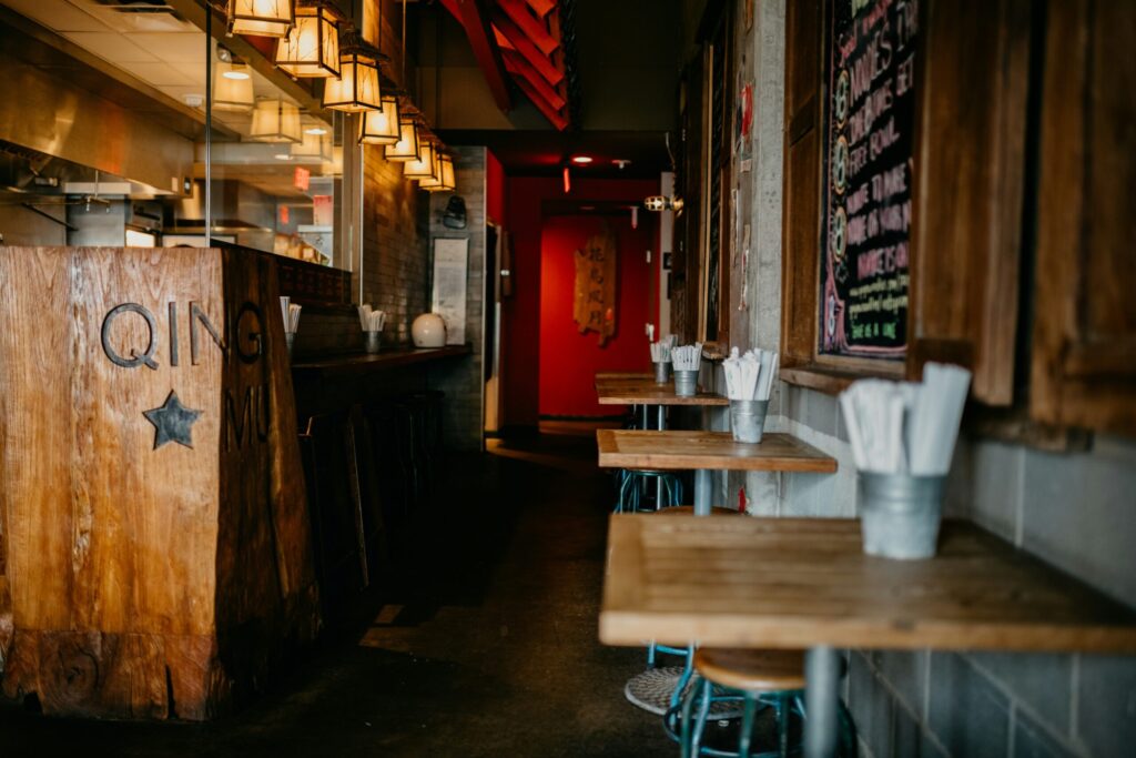 Narrow restaurant interior with wooden bar counter, small wood tables lined along the wall, hanging lantern-style lights, and chopsticks set on each table.