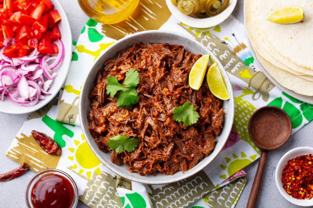 Bowl of shredded beef topped with cilantro and lime wedges, surrounded by tortillas, chopped onions, tomatoes, sauces, and spices on a patterned table.
