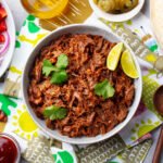 Bowl of shredded beef topped with cilantro and lime wedges, surrounded by tortillas, chopped onions, tomatoes, sauces, and spices on a patterned table.
