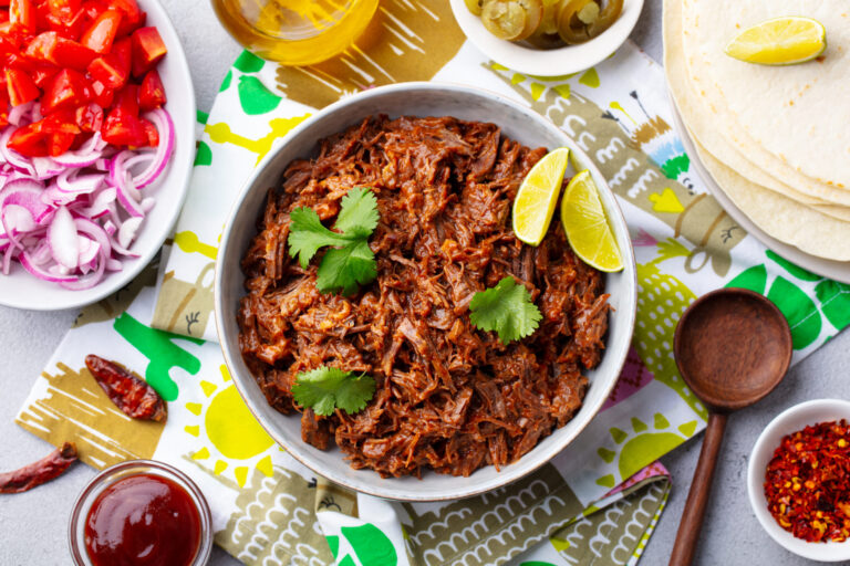 Bowl of shredded beef topped with cilantro and lime wedges, surrounded by tortillas, chopped onions, tomatoes, sauces, and spices on a patterned table.