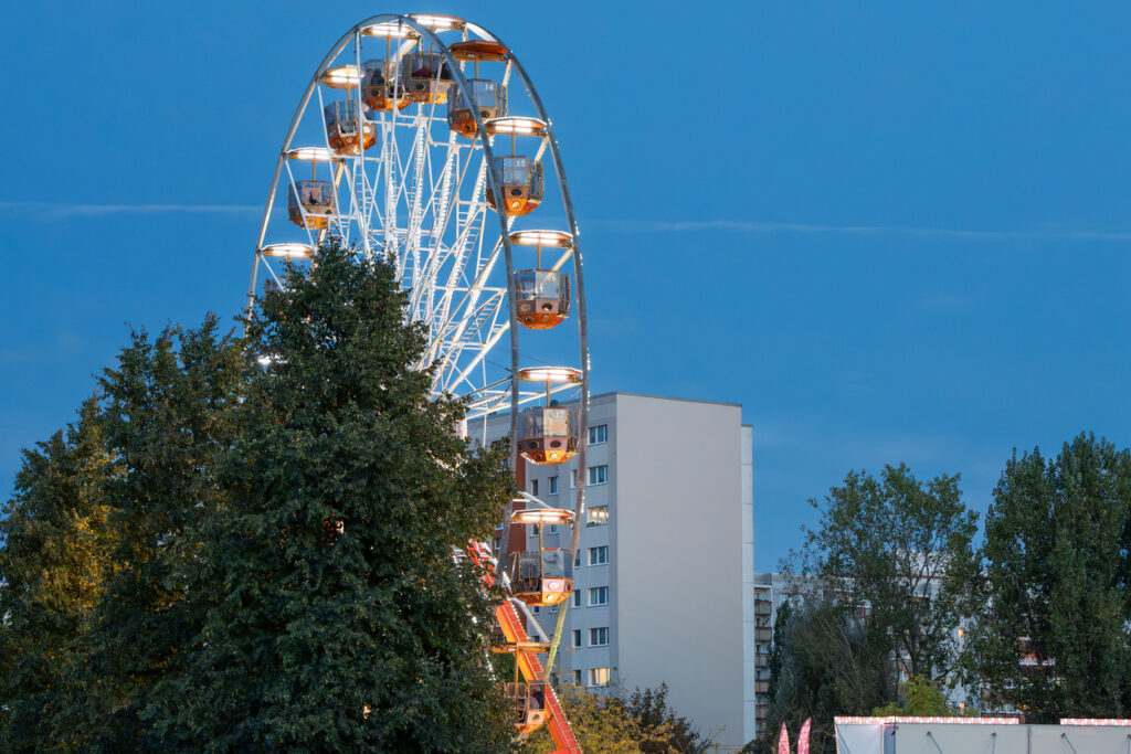 Ferris wheel illuminated at dusk behind trees and apartment buildings