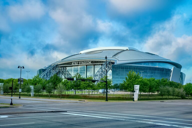 Modern stadium exterior with curved glass and metal facade, viewed from a street intersection under a partly cloudy blue sky.