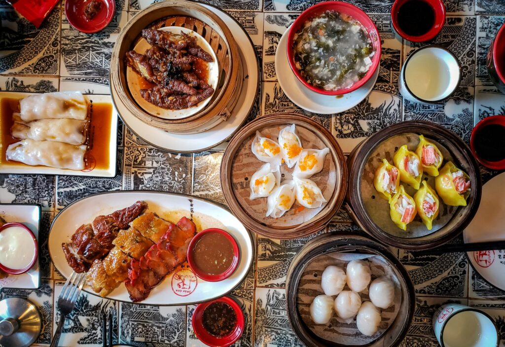 Overhead view of a dim sum spread with steamed dumplings, buns, rice rolls, soups, and sauces arranged on a patterned tablecloth.