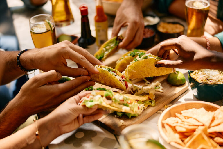 People sharing tacos around a table with drinks, chips, and dipping sauces.