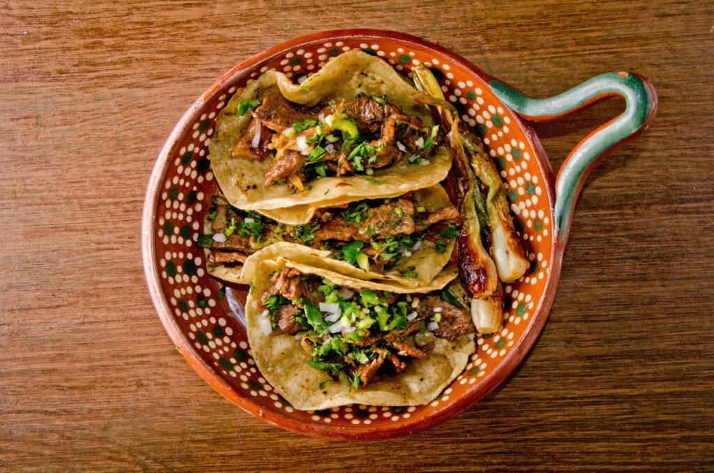 Three beef tacos topped with chopped cilantro and onions, served in a decorative ceramic bowl on a wooden table.
