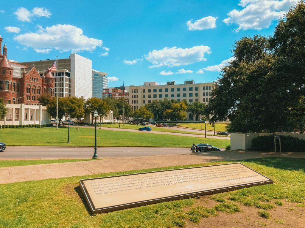 View of Dealey Plaza in Dallas with grassy lawn, historic buildings, and a plaque in the foreground.