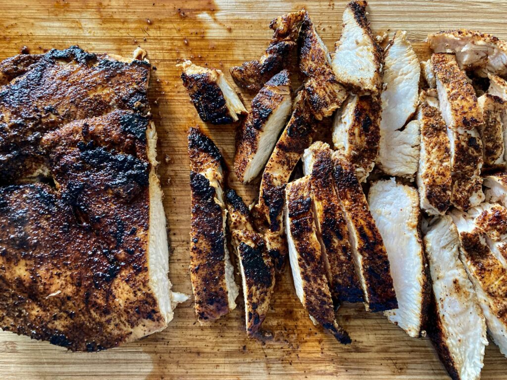 Sliced grilled chicken breast with charred seasoning on a wooden cutting board.